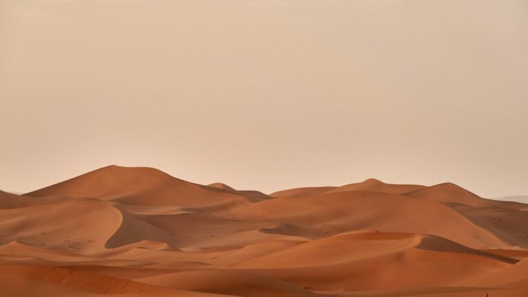 Wind lifting sand from the crest of a desert dune under a pale sky